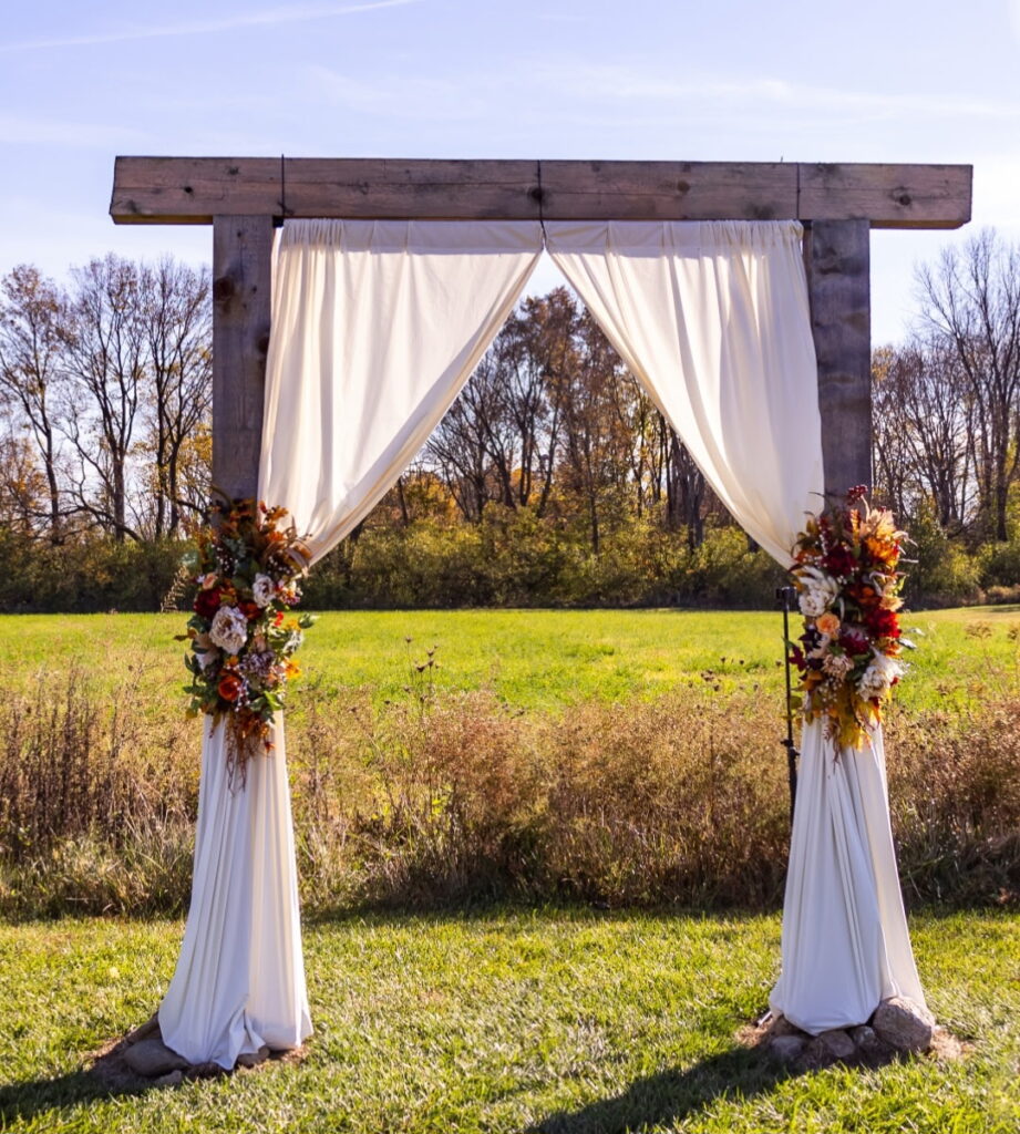 Rustic wooden wedding arch with white drapes and fall florals in a field